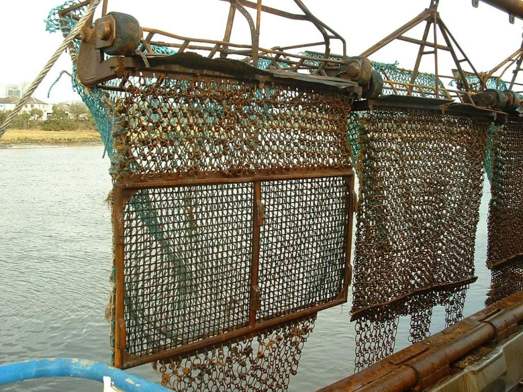 Three dredges mid-air on the side of vessel. The dredges are square shaped metal frames with netting on the top half and a metal grid on the bottom