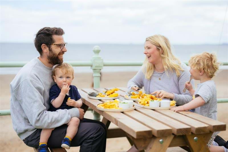 A family eating a fish and chip takeaway on a picnic bench beside the beach