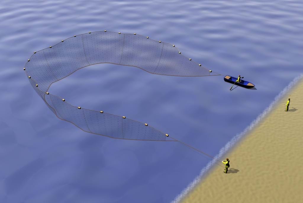 Man standing on beach holding one end of net, whilst a small boat completes a semi-circle, paying out the net as it does.