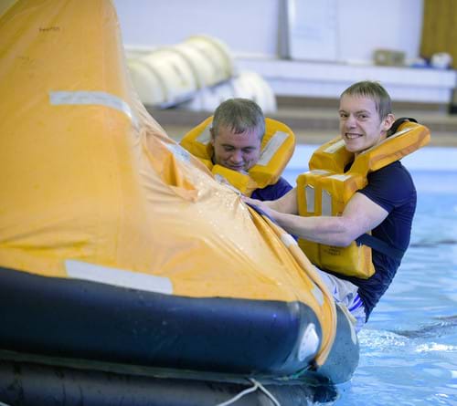 Two men taking part in mandatory sea survival training. They are in a swimming pool holding on to a life raft as part of the training.