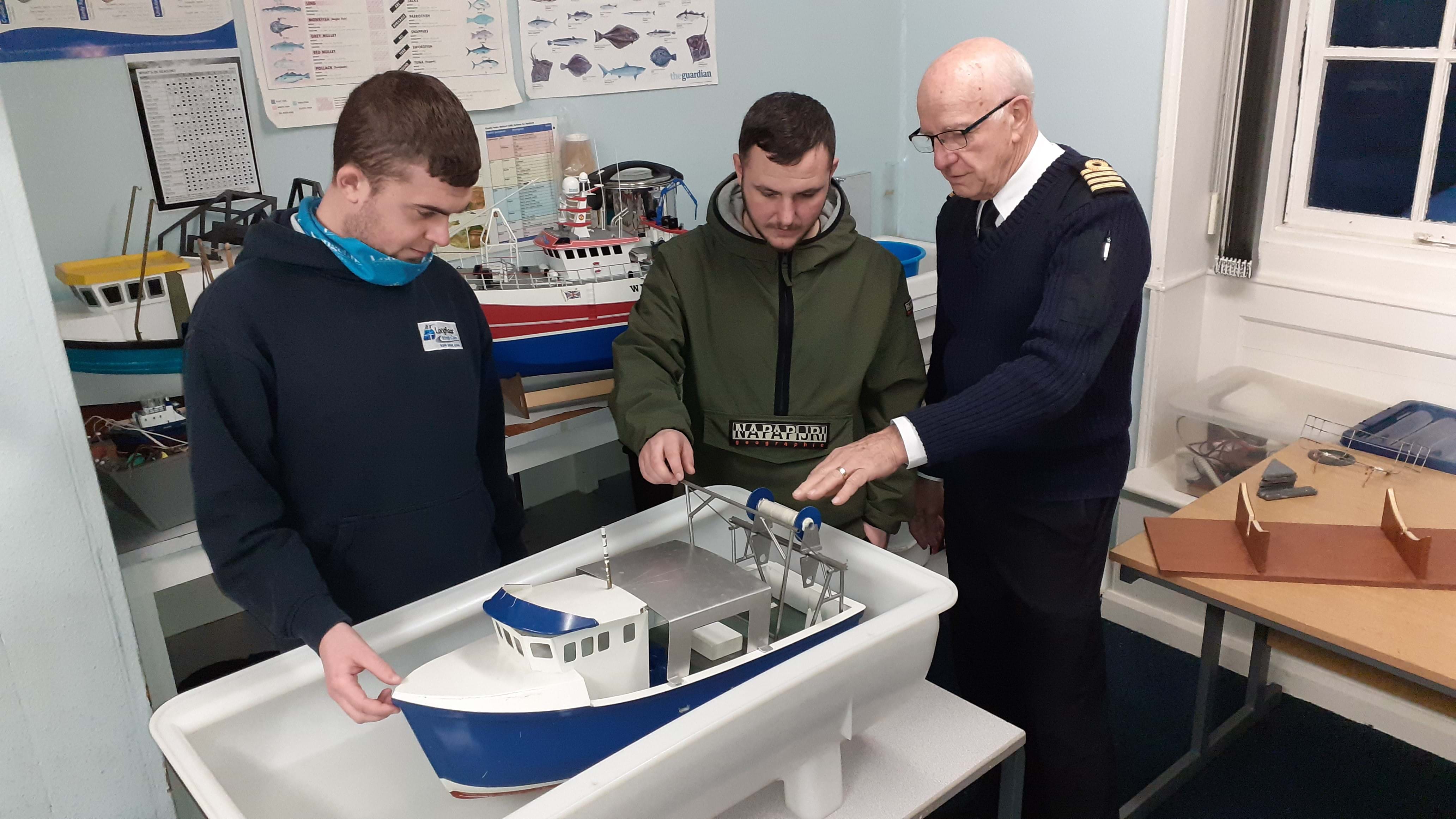 Photo shows two men being shown a model fishing vessel by a trainer in a classroom.