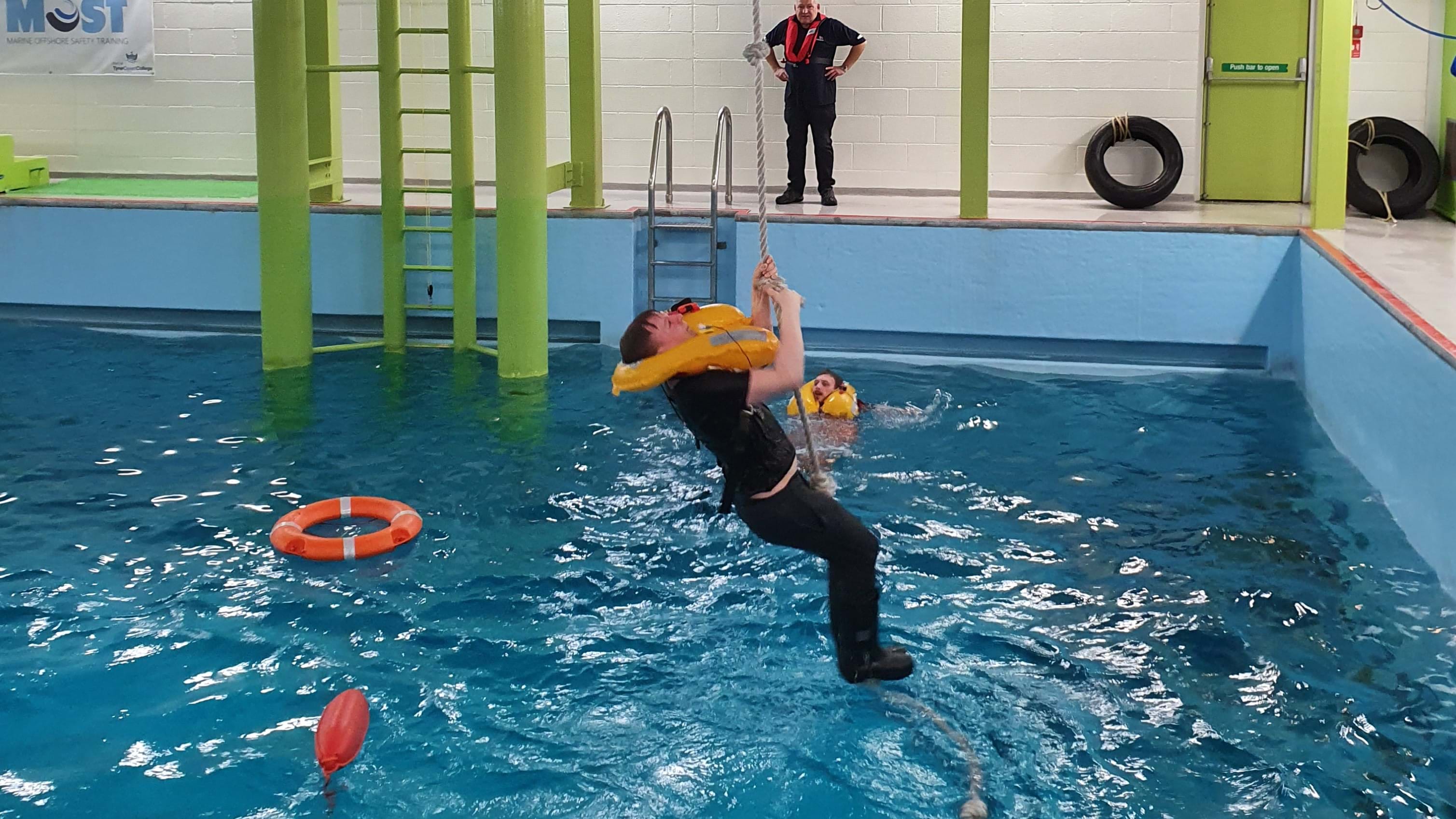 A man is in a training pool being lifted by a winch a rope to simulate a helicopter rescue.