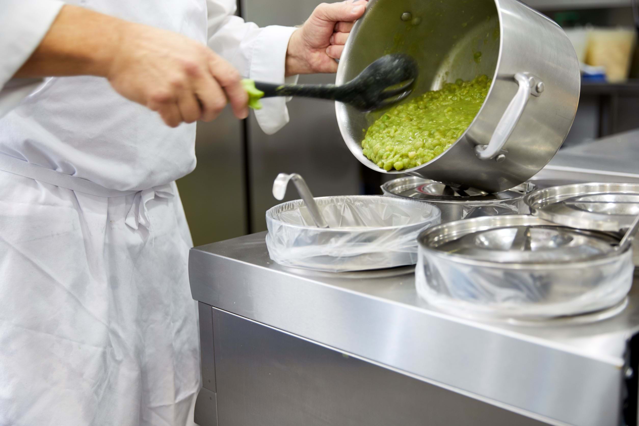 Photo shows someone pouring mushy peas from a pot in to a bain marie for serving.